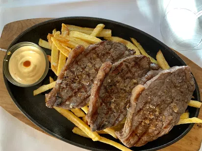 Plato de carne a la parrilla con patatas fritas y salsa en un recipiente peque&ntilde;o, servido en una tabla de madera.