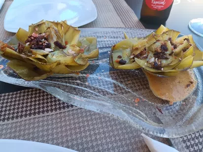 Dos alcachofas asadas servidas en un plato de cristal, decoradas con trozos crujientes y acompa&ntilde;adas de una salsa cremosa.